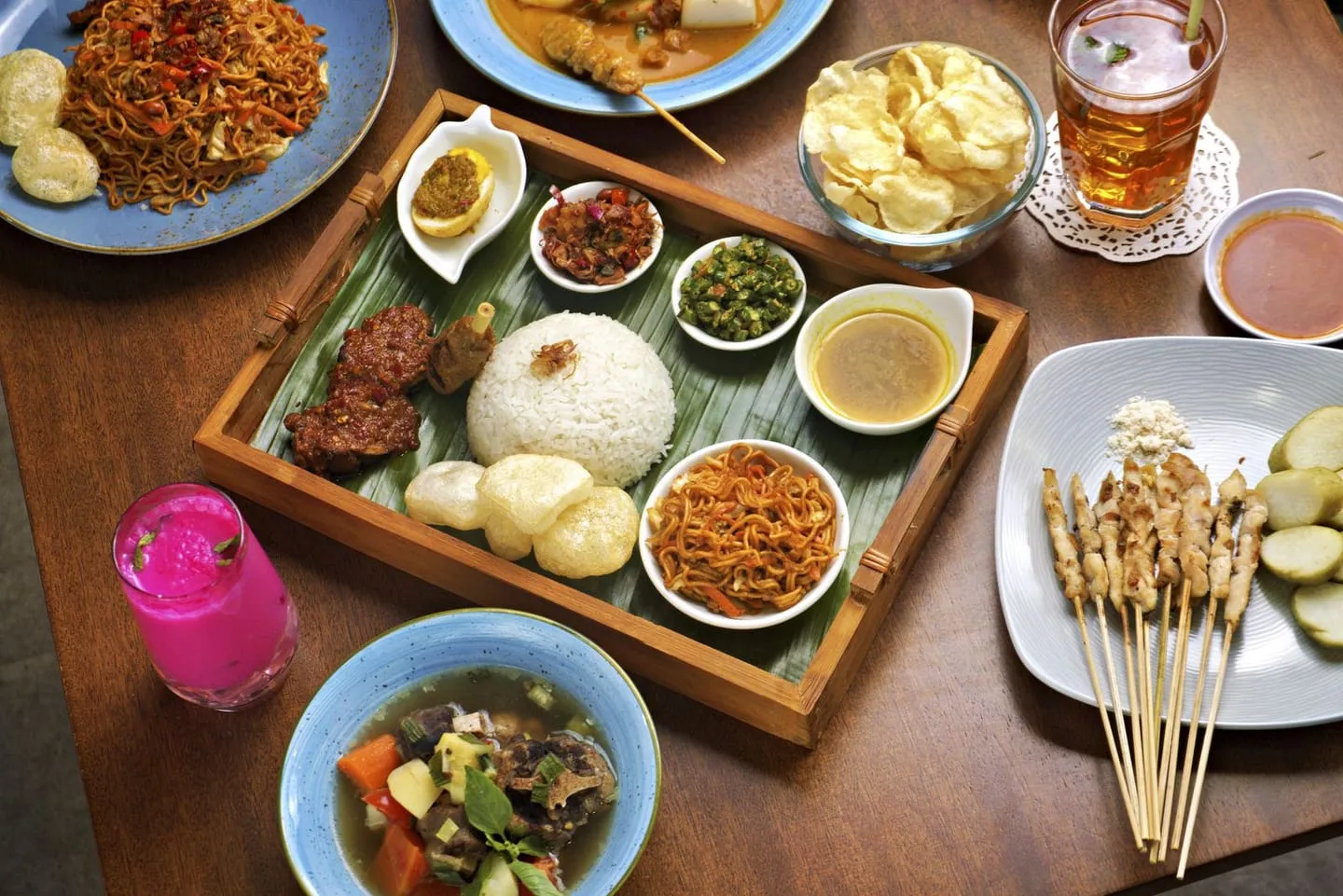 Hidangan sate, nasi campur, dan aneka lauk di atas meja kayu