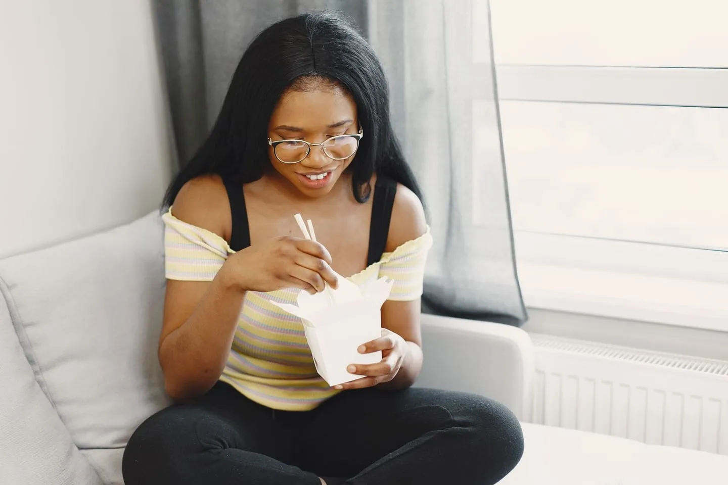 A young woman eating takeout at home by a window